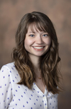 Photo of young woman with brown hair and bangs wearing a white shirt with tiny blue pattern, 2018.