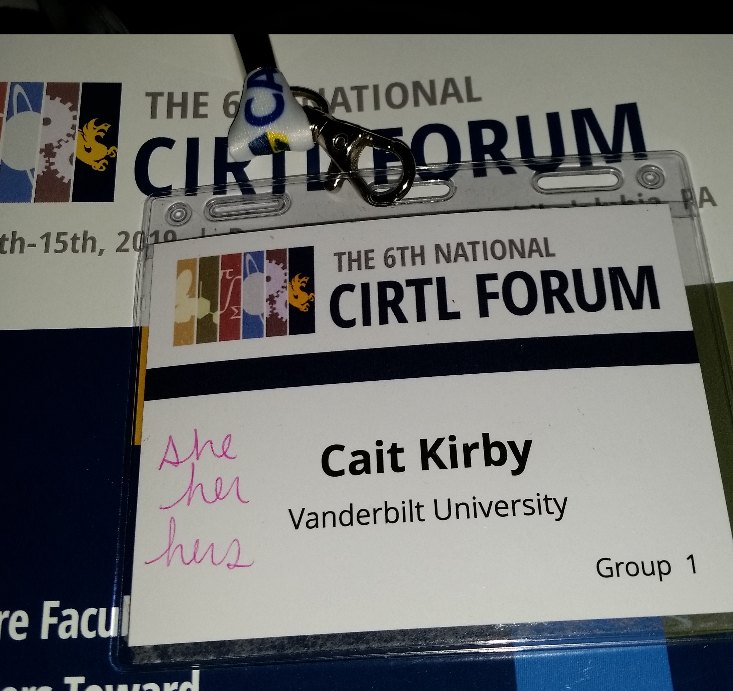 Photo of black text on white background printed nametag that says: Cait Kirby Vanderbilt University. Handwritten in pink it says: she/her/hers.